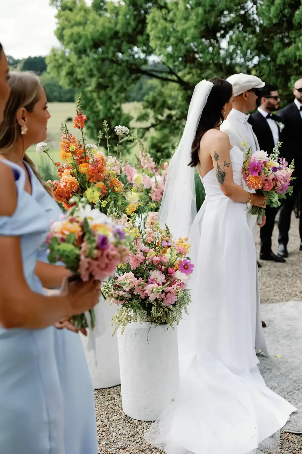 A group of people dressed in wedding attire standing outdoors with floral arrangements and greenery in the background.