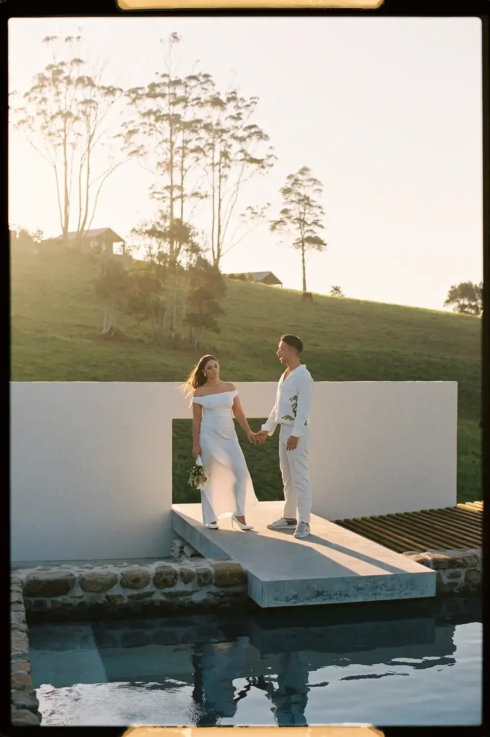 A couple dressed in white, holding hands and standing on a small bridge over water at sunset. The woman wears a white off-shoulder dress holding a bouquet of flowers, and the man wears a white suit with floral designs. The background features a grass