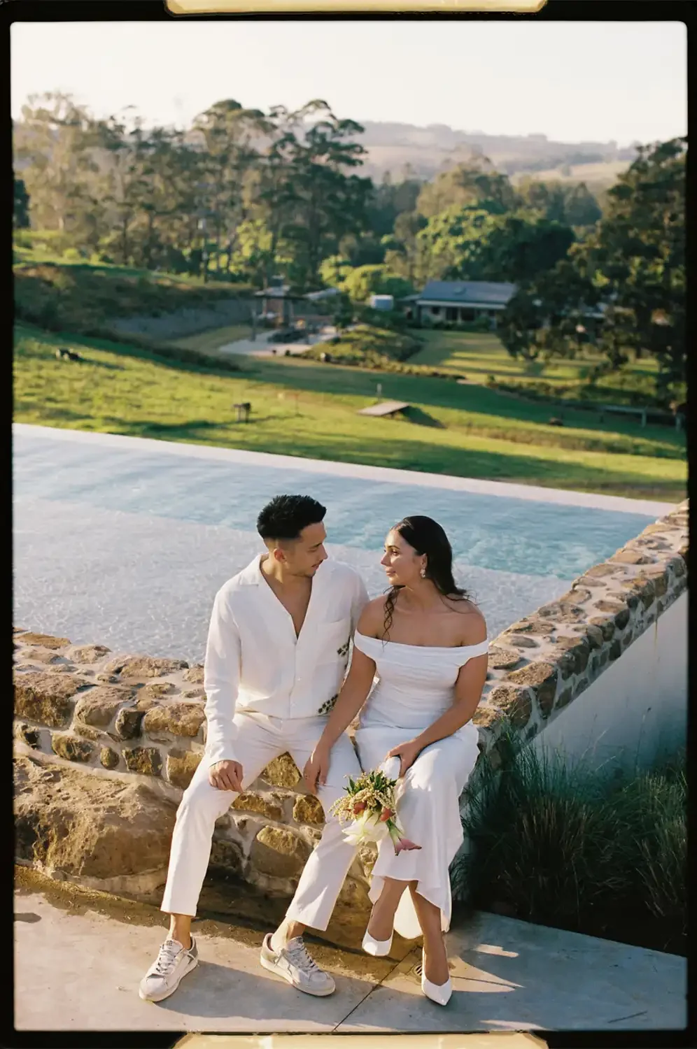 A couple dressed in white sitting on a stone wall near a pool with a green landscape and trees in the background.