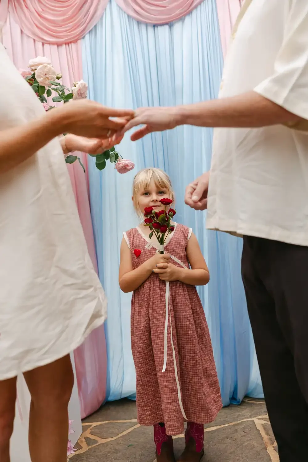 A young girl in a pink dress holding a bouquet of red roses in a decorated setting with pastel-colored drapes and pink roses, participating in a ceremony or event.