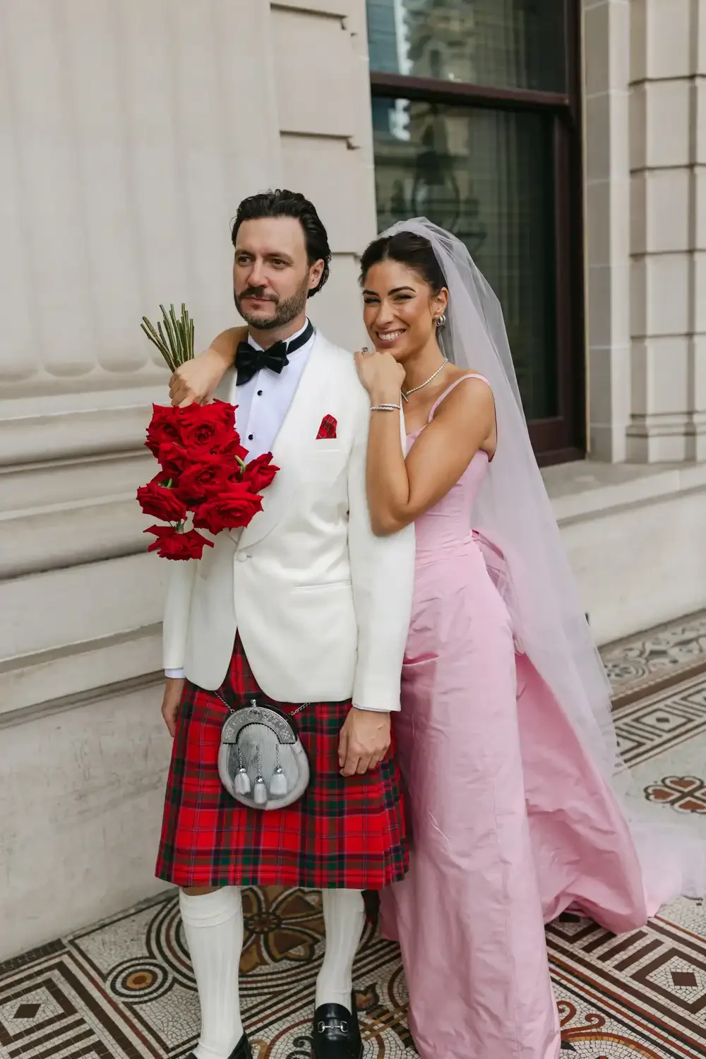 A bride and groom standing outside a building, with the groom holding a bouquet of red roses and dressed in a tuxedo with a kilt, and the bride wearing a pink wedding gown with a veil, smiling and embracing each other.
