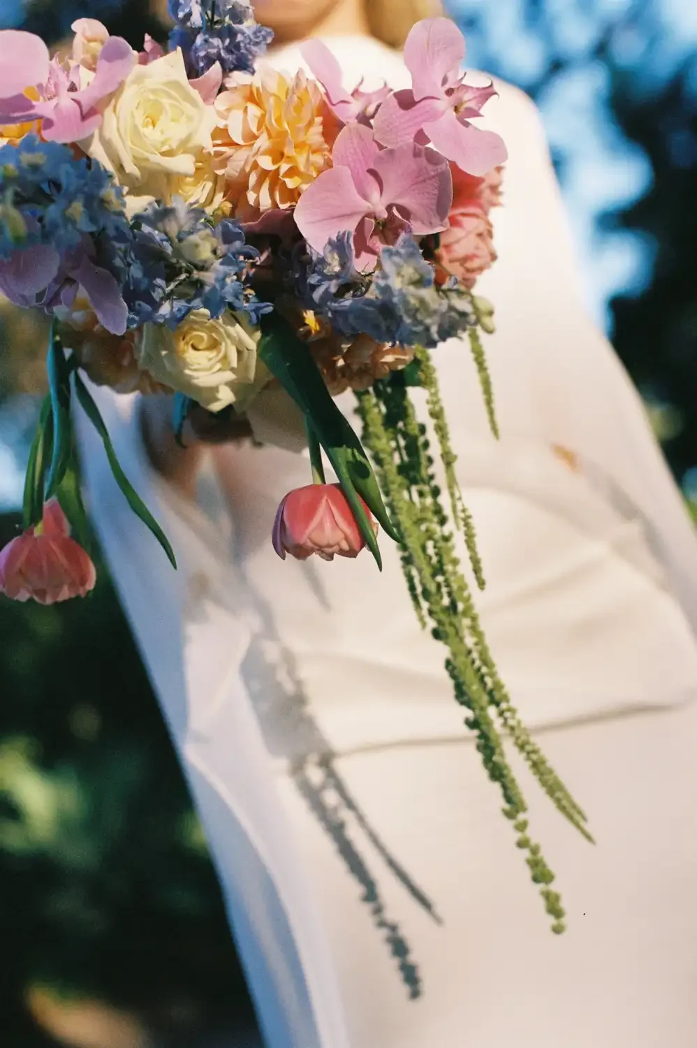 A person holding a large bouquet of colorful flowers, including roses, orchids, and tulips, outdoors in natural light.
