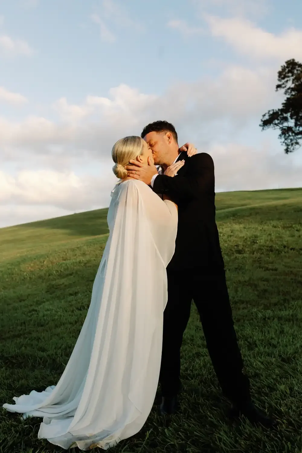 A couple dressed in wedding attire sharing a kiss outdoors on a grassy hill under a partly cloudy sky.
