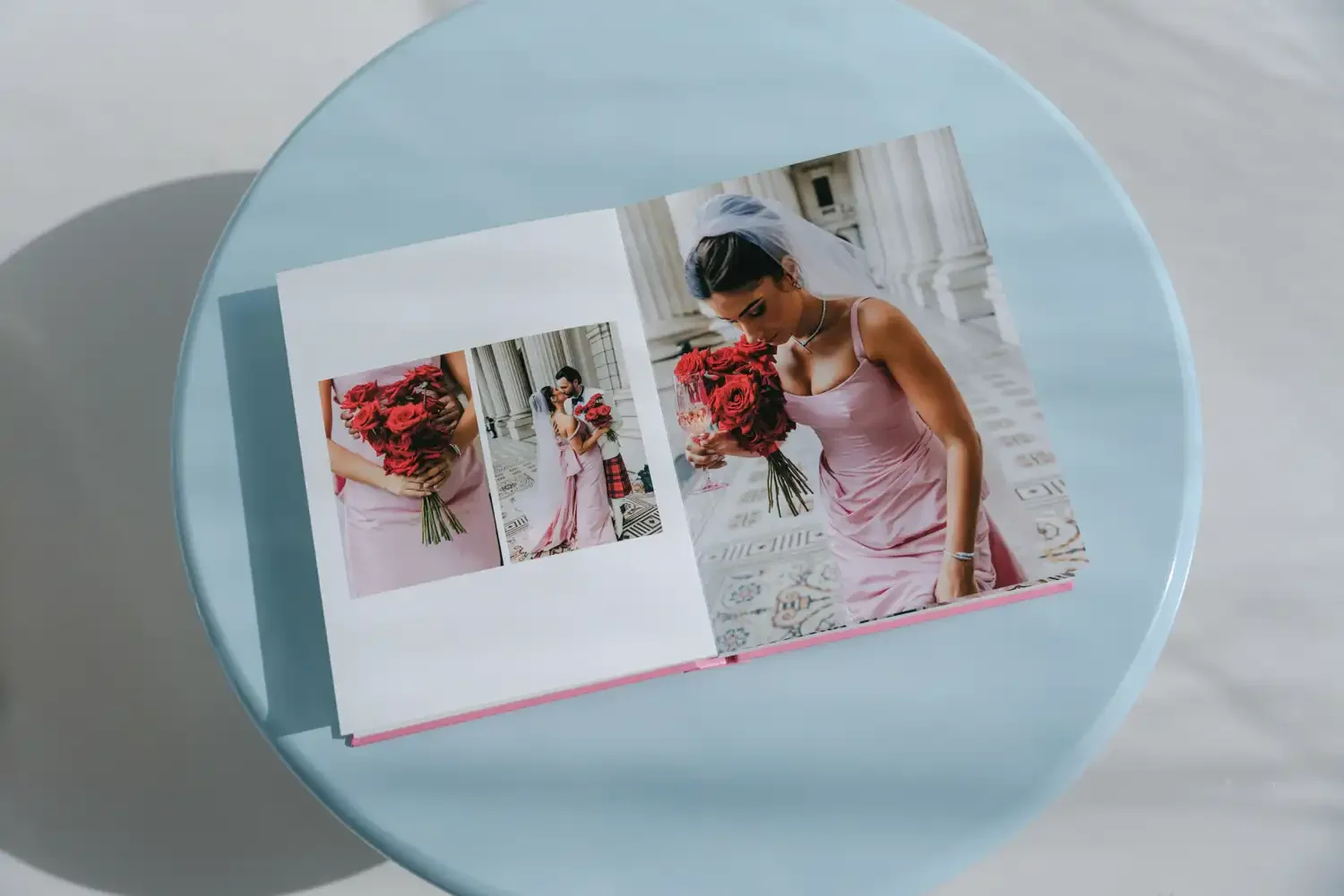 Open photo album on a light blue table showing two pictures of a woman in a pink satin dress holding a bouquet of red roses, standing on a patterned floor with white columns in the background.