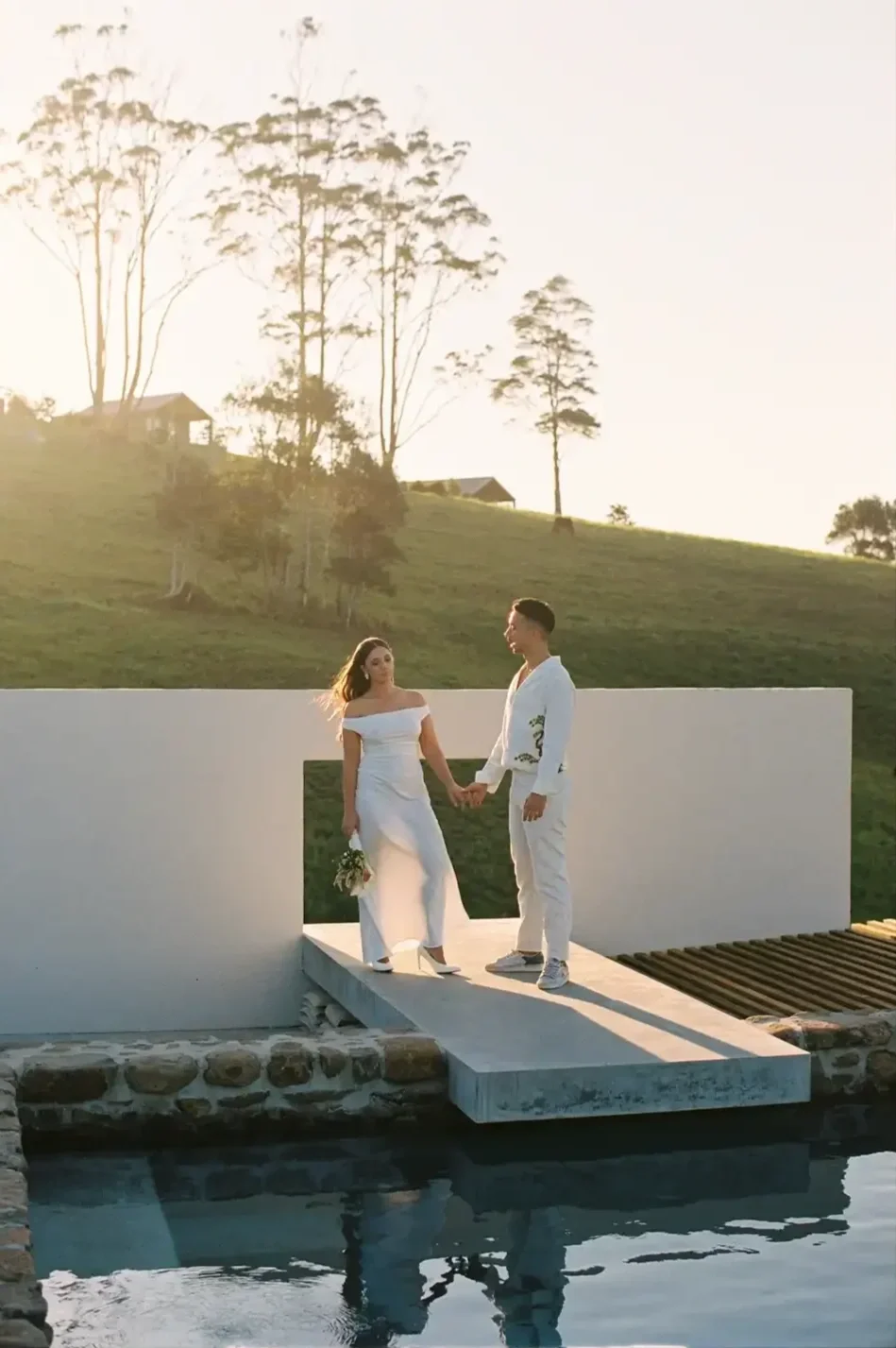 A couple dressed in white wedding attire holding hands on a small bridge by a reflecting pool, with a grassy hillside and trees in the background during sunset.