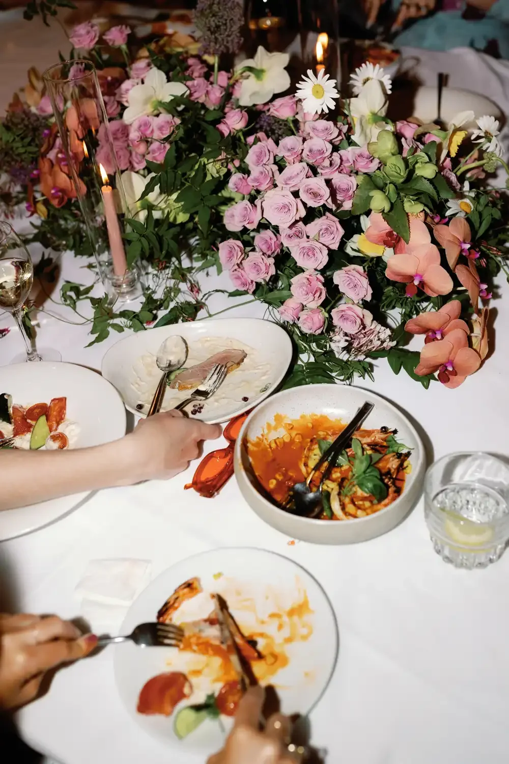 A dinner table with floral centerpieces featuring pink roses, white daisies, and other flowers. There are plates with partially eaten food, glasses of water with lemon, and candles in glass holders.