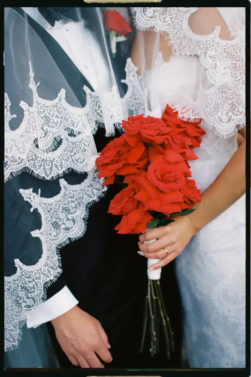 Close-up of a bride holding a bouquet of red roses, standing next to a groom dressed in a tuxedo with a white shirt and bow tie, both wearing lace garments.