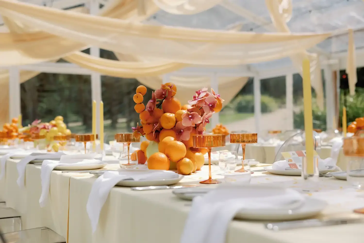 Elegant outdoor table set for a celebration with a centerpiece of oranges and pink flowers, under a white canopy, with greenery in the background.