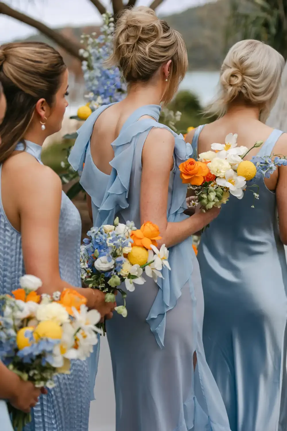 Women in light blue dresses holding bouquets of white, orange, yellow, and blue flowers outdoors