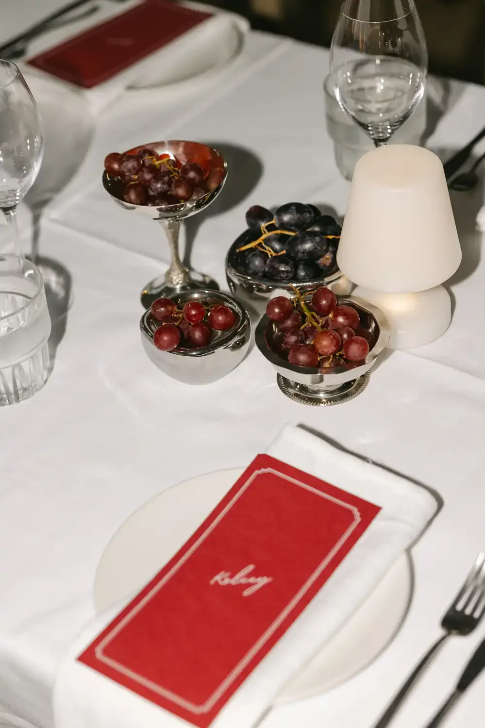 A formal dining table setup with a white tablecloth, glasses, a white lamp, a small bowl of red grapes, a larger bowl of black grapes, and a menu with a red cover that says "today."