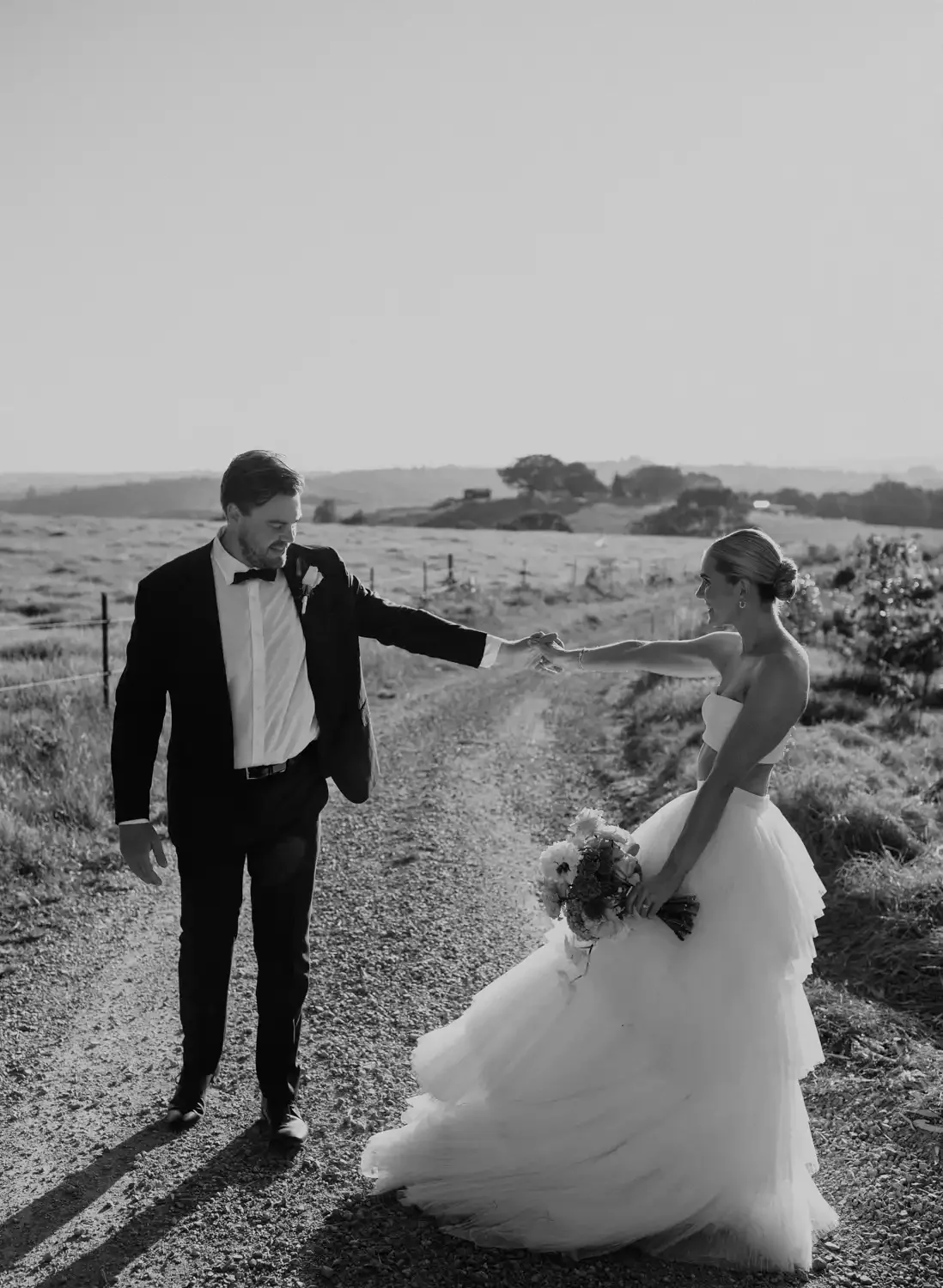 Black and white photo of a bride and groom walking on a rural dirt path, holding hands and smiling, with open fields and distant trees in the background.