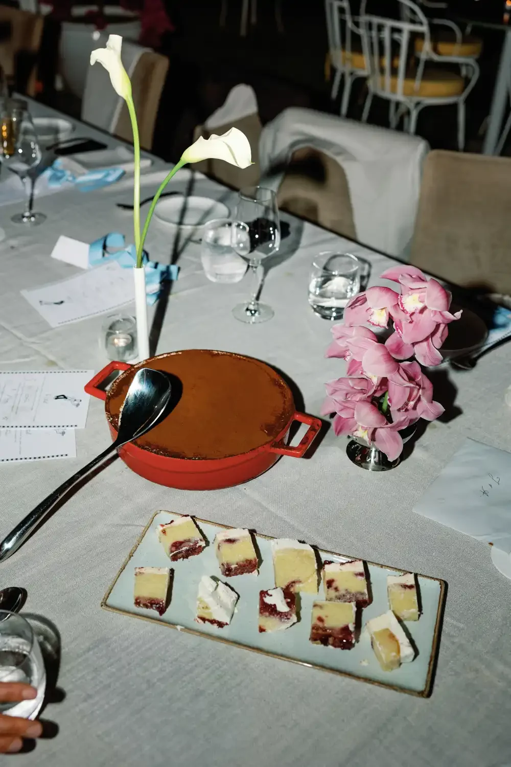 Table set with flowers, glassware, a chocolate dessert in a red dish, and sliced cheesecake on a white platter at an elegant restaurant.