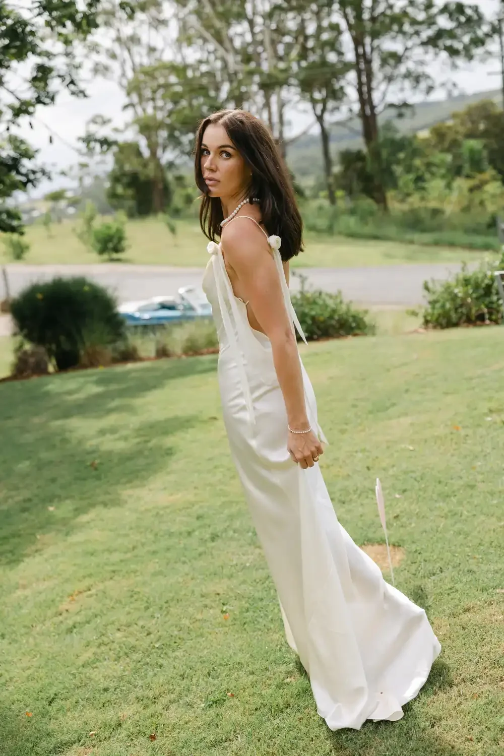 A woman in a white dress standing on a grassy lawn with trees and a road in the background.