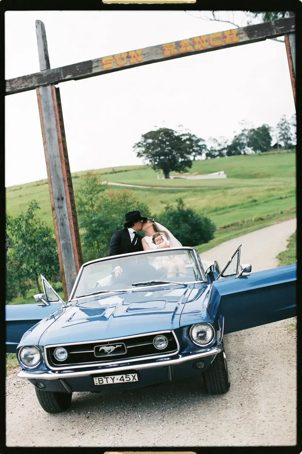 A couple in vintage black and white wedding attire kissing inside a blue classic Ford Mustang convertible, with a woman holding a small child, parked on a country road, green hills and trees in the background.
