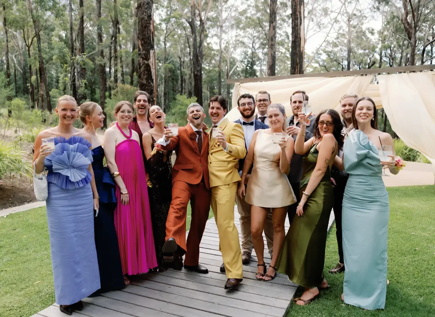 Group of people at a celebration outdoors, dressed in formal attire, holding drinks, standing on a wooden pathway with trees in the background.