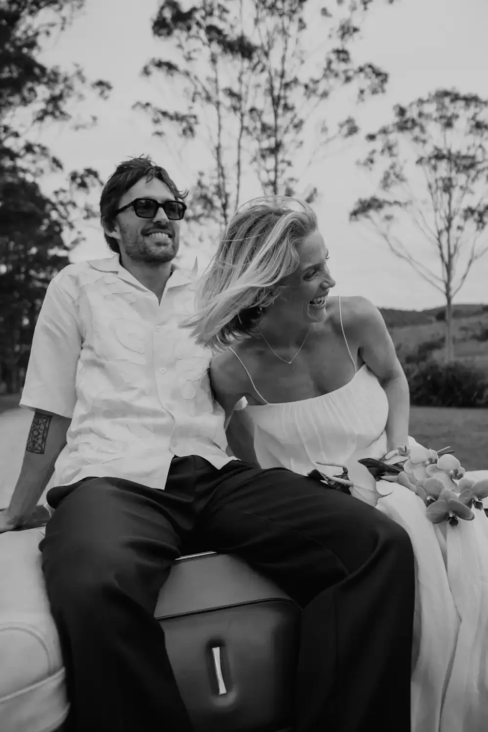 A black-and-white photo of a man and woman laughing and sitting together outdoors, with trees and a field in the background. The woman holds a bouquet of flowers.