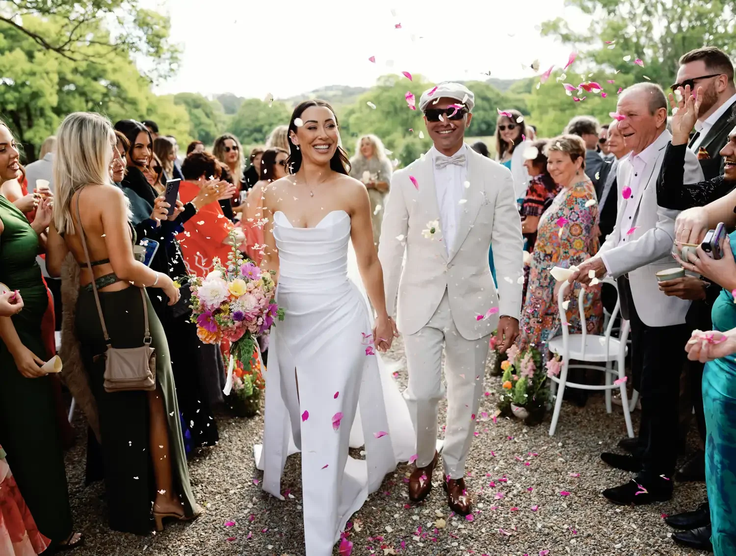 A newly married couple walking hand in hand through a celebration crowd, with pink flower petals falling, outdoors with green trees in the background. The bride is in a white strapless gown holding a bouquet, and the groom is in a white suit with sun