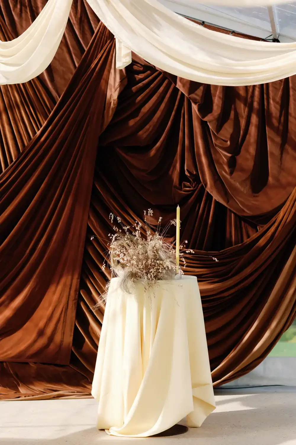 A tall white candle and dried floral arrangement on a cream-colored draped table in front of brown velvet curtains with white fabric accents.