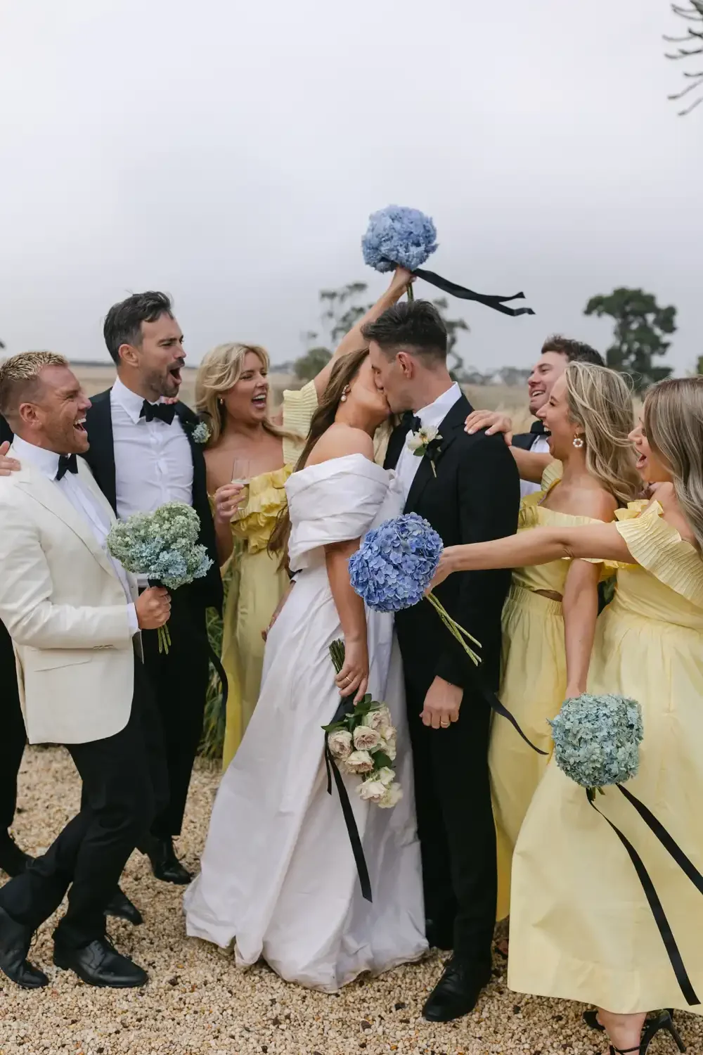 A wedding celebration with the bride and groom kissing while surrounded by friends in colorful attire, holding bouquets of flowers, outdoors on a cloudy day.