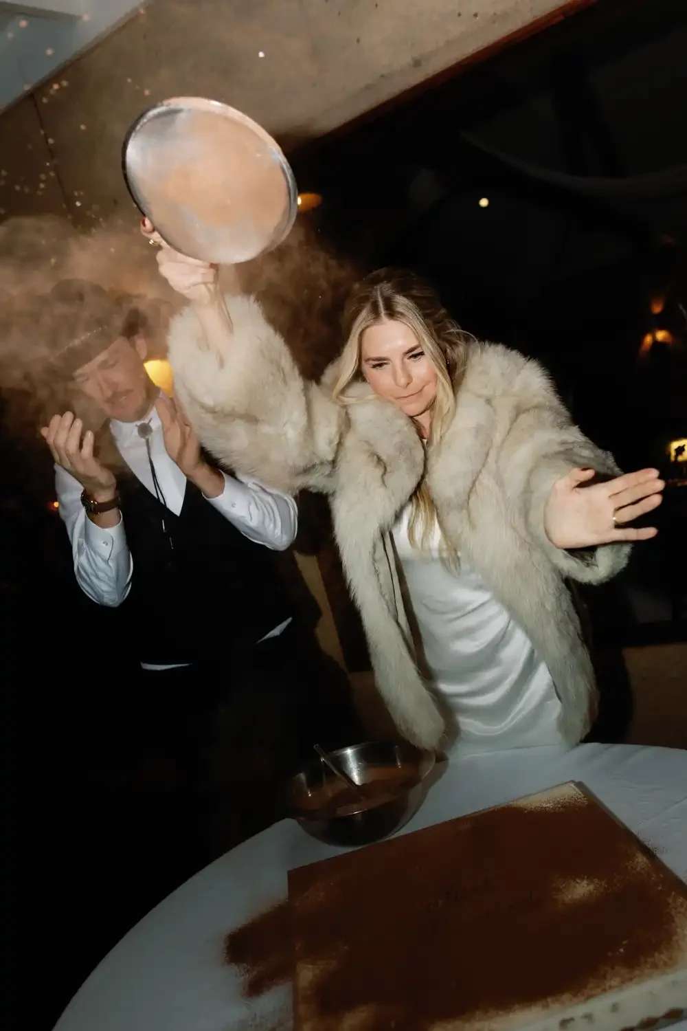 A woman in a faux fur coat and a man in a dress shirt and vest celebrate around a cake with cocoa powder, with the woman playfully shielding her face from the man who is tossing flour or powdered sugar.