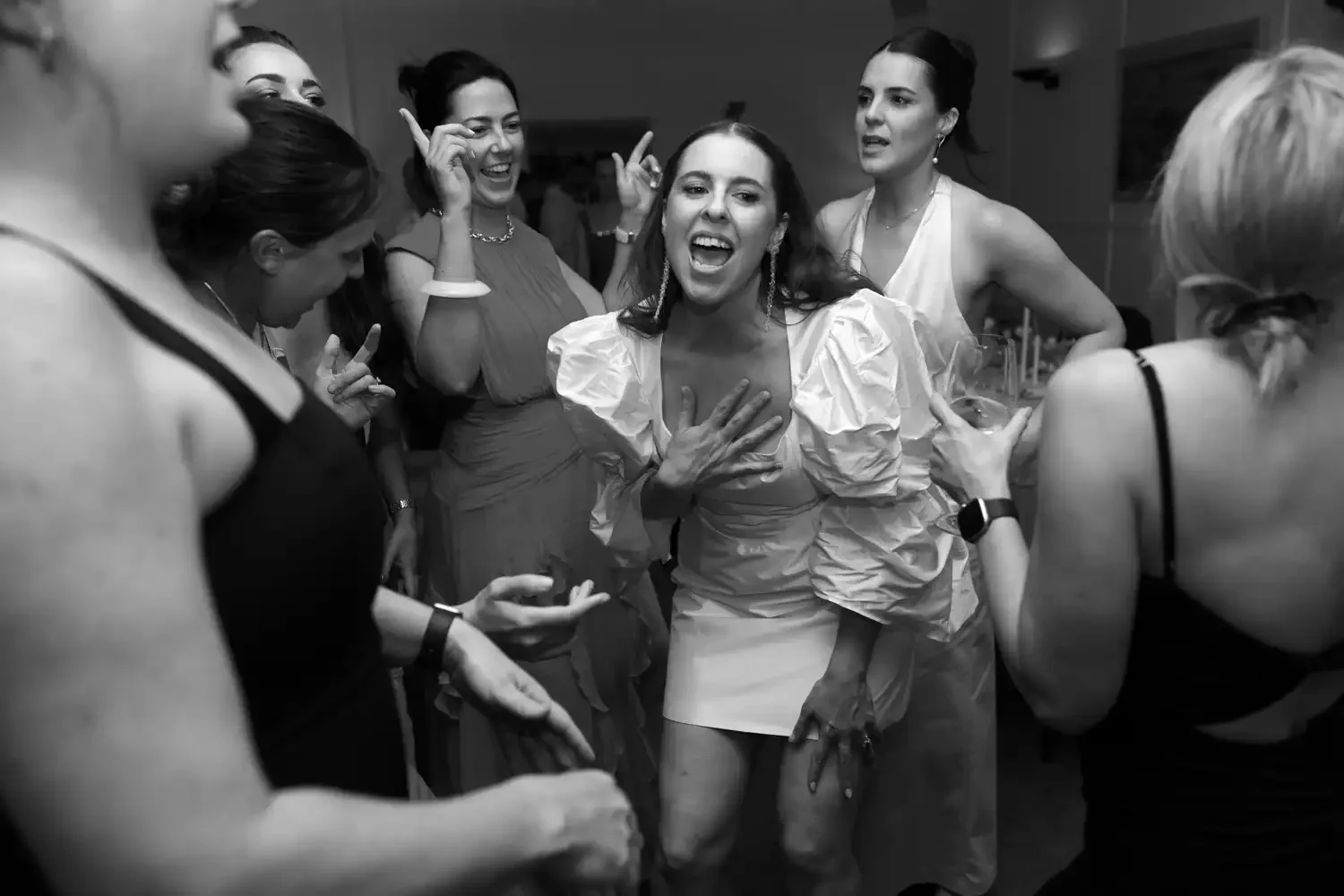 Women dancing and celebrating at a party, with one woman in the center holding her chest and singing or shouting joyfully.