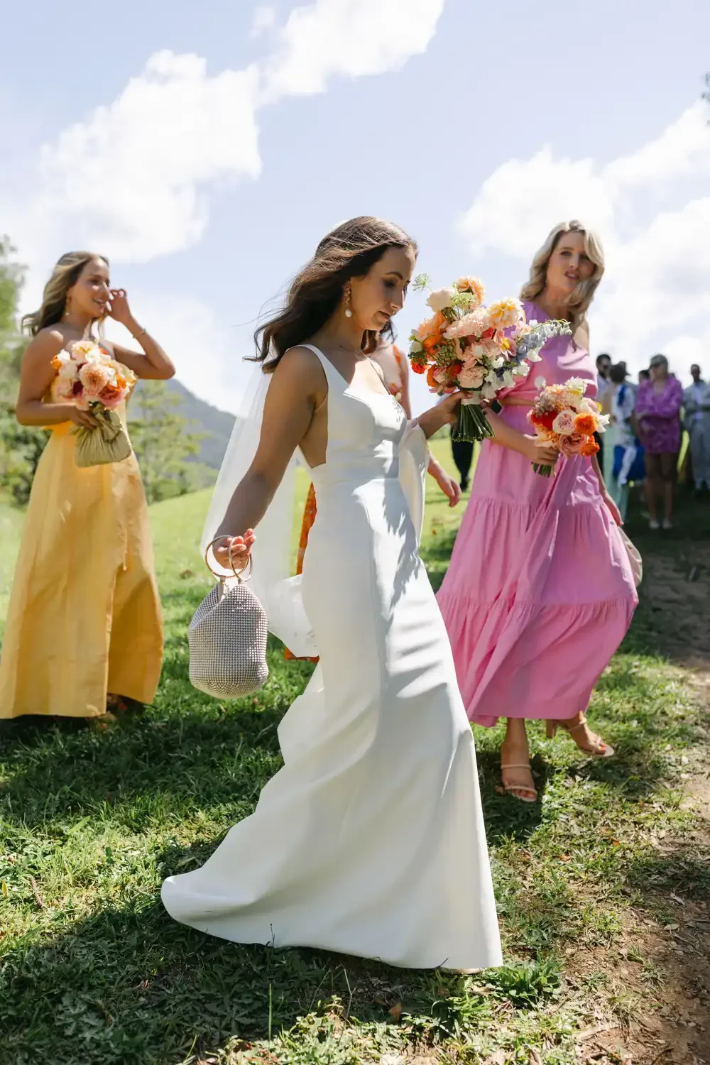 Bridal party walking outdoors with bride in white dress holding a bouquet, surrounded by bridesmaids in colorful dresses, holding bouquets of flowers, on a sunny day with blue sky and clouds.