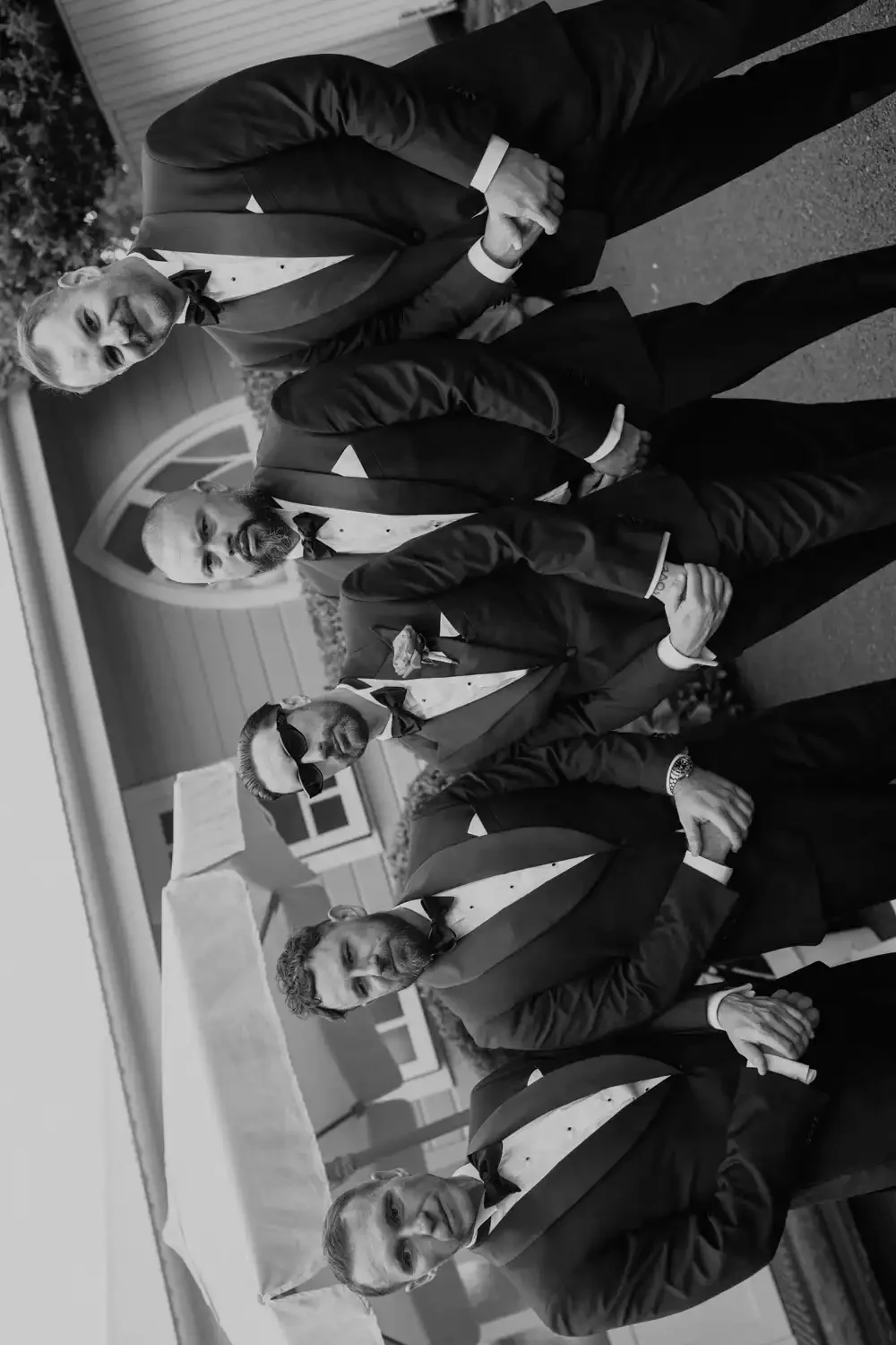 A group of six men dressed in tuxedos, standing outdoors in front of a house, posing for a photo with some holding an umbrella.