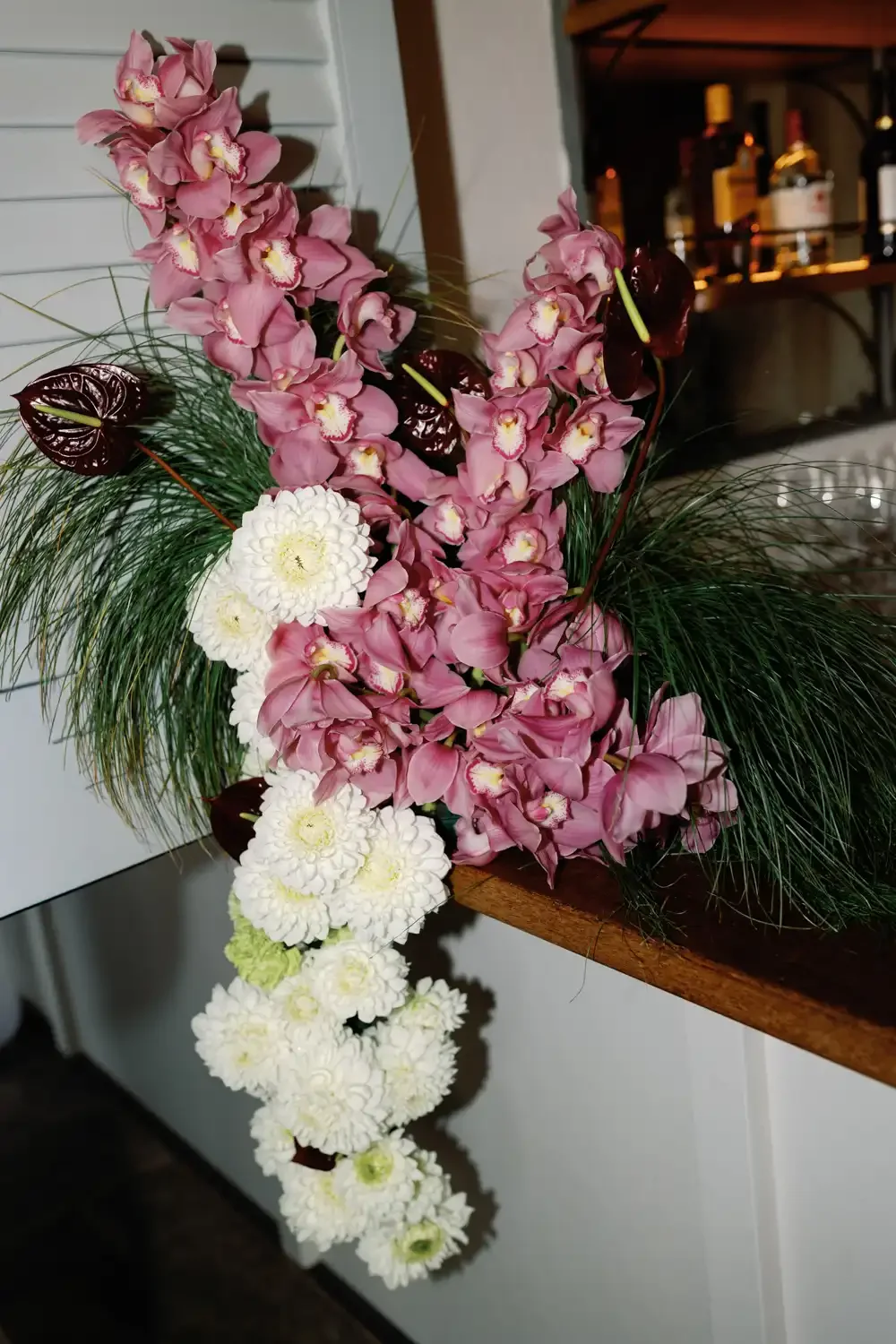 A floral arrangement with cascading white chrysanthemums, pink orchids, dark red anthuriums, and greenery on a wooden shelf.