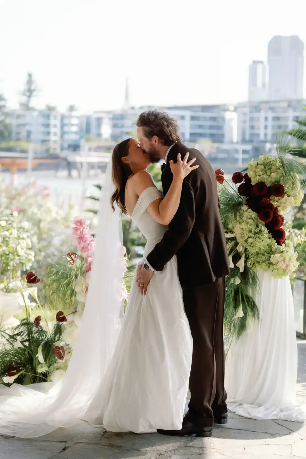 A bride and groom sharing a kiss during their wedding ceremony, standing in front of floral decorations.