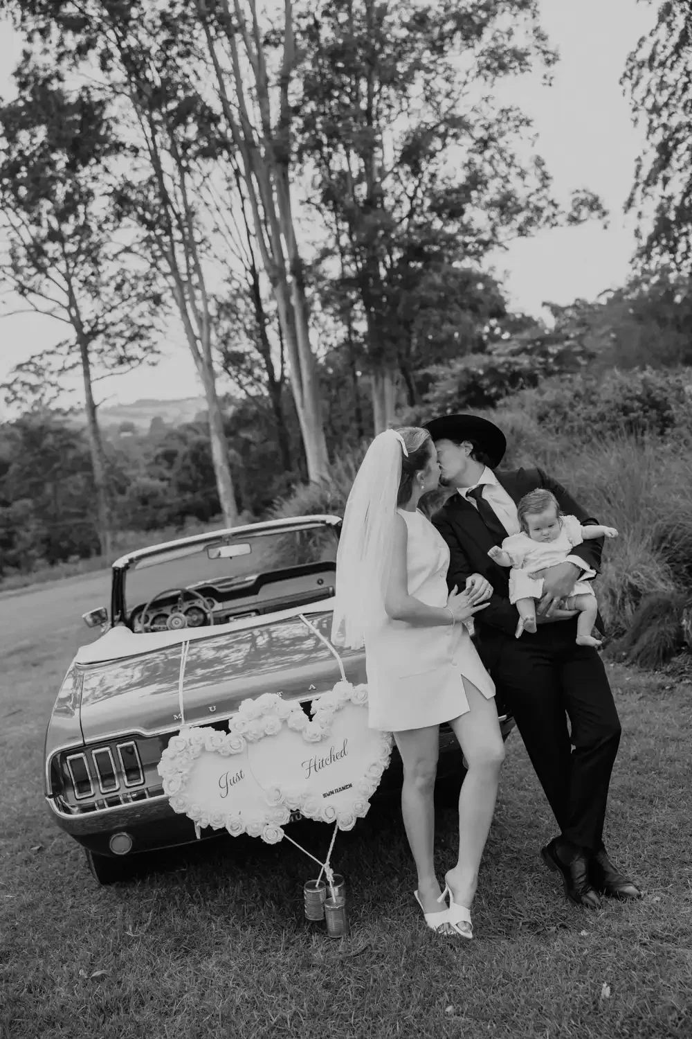 A black and white photo of a newlywed couple kissing outdoors next to a vintage convertible car decorated with a sign that reads "Just Married." The groom, dressed in a suit and cowboy hat, holds a baby girl, while the bride, in a sleeveless dress an