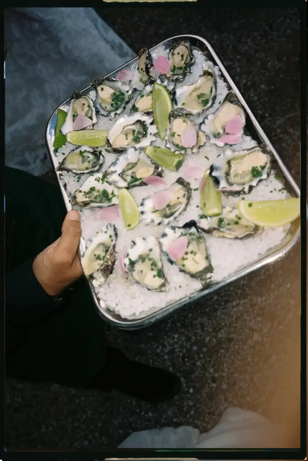 A person holding a tray of raw oysters garnished with lime wedges, pink flower petals, chopped chives, and served on a bed of ice.