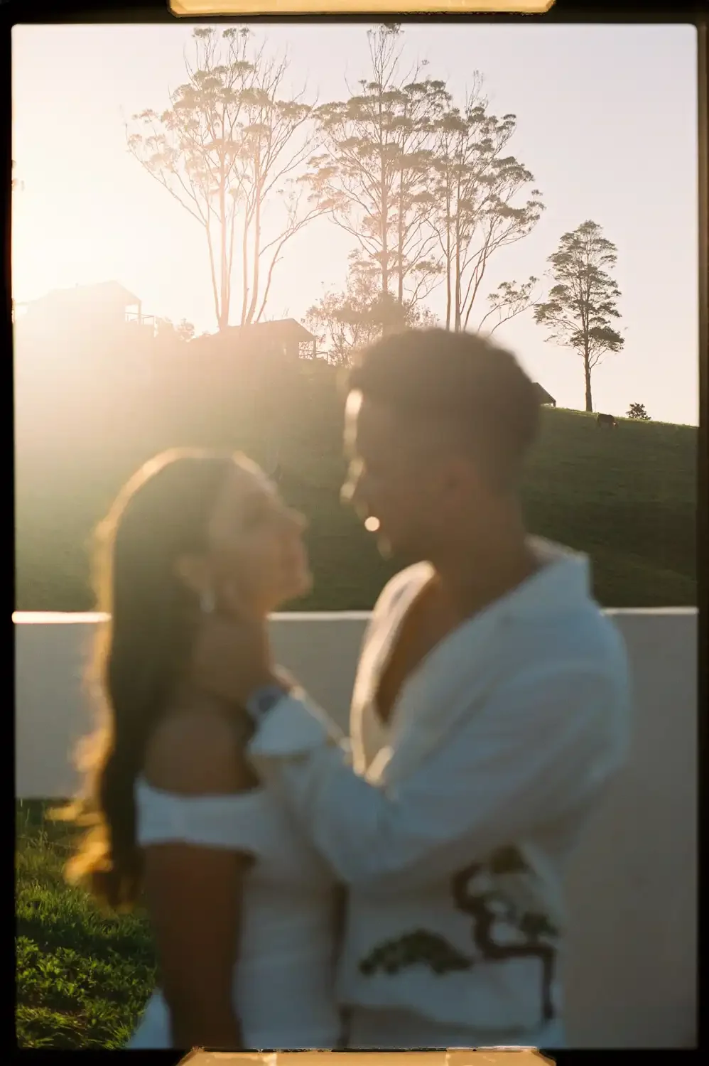 A blurred photo of a couple facing each other outdoors at sunset, with trees and houses in the background.
