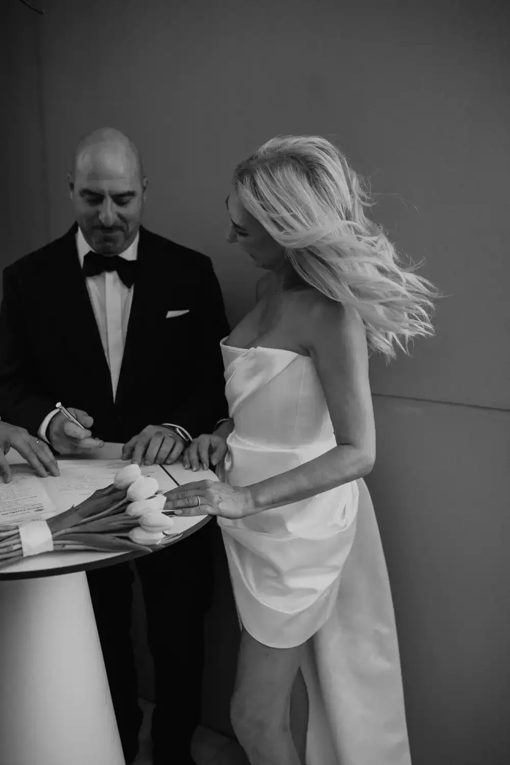 A black-and-white photo of a wedding ceremony with a bride in a strapless dress and a groom in a tuxedo signing documents at a table decorated with spring flowers.