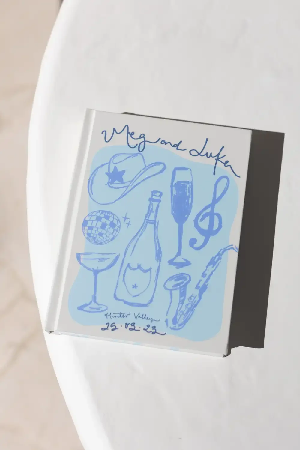 Invitation with hand-drawn illustrations of a cowboy hat, disco ball, wine glass, champagne bottle, treble clef, saxophone, and wine glass, with handwritten text 'Megan and Duke' and event details 'Hunter Valley 25.02.23'.