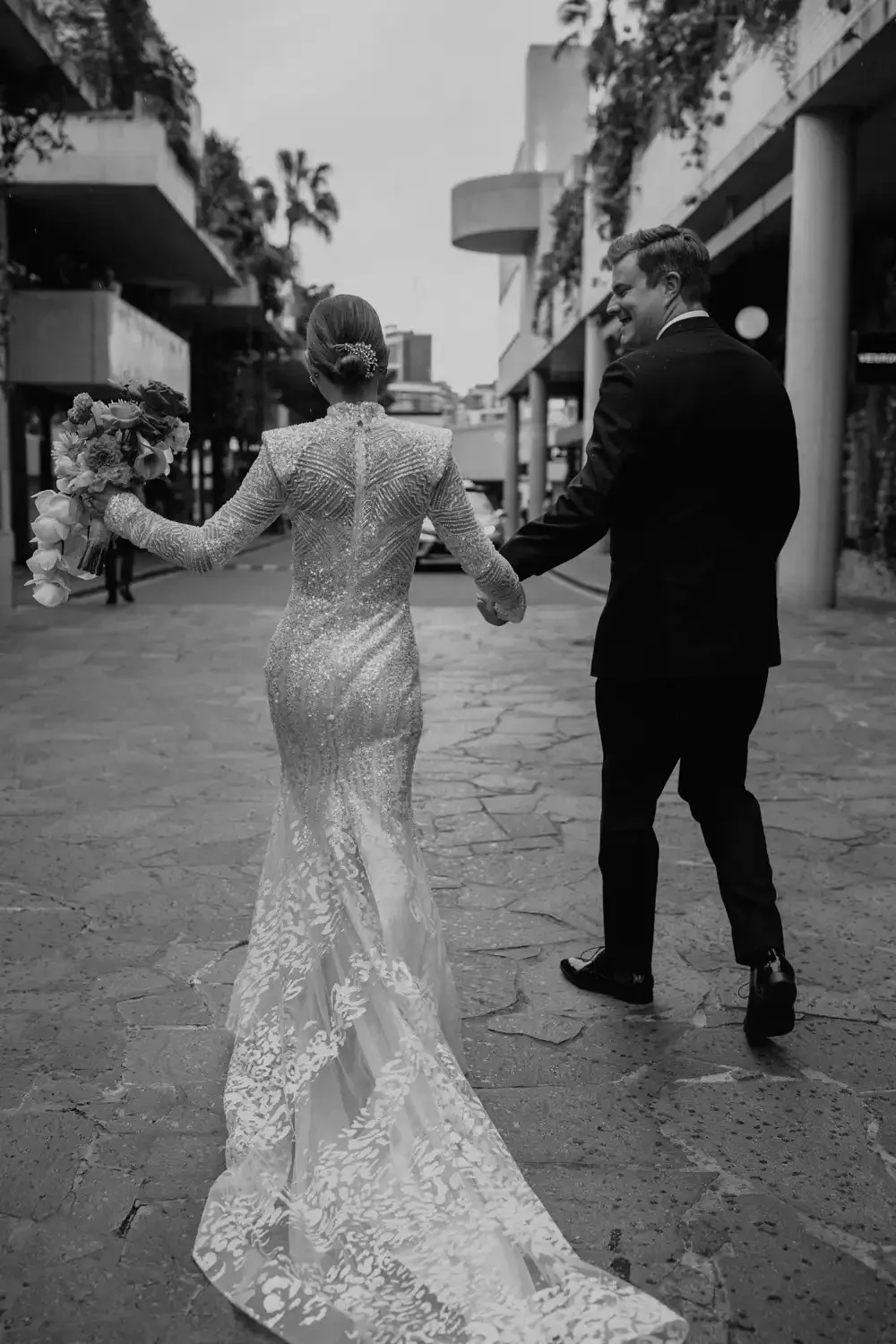 A bride and groom walk hand in hand on a city street, smiling and holding a bouquet, with the bride in a detailed lace wedding gown and the groom in a dark suit.