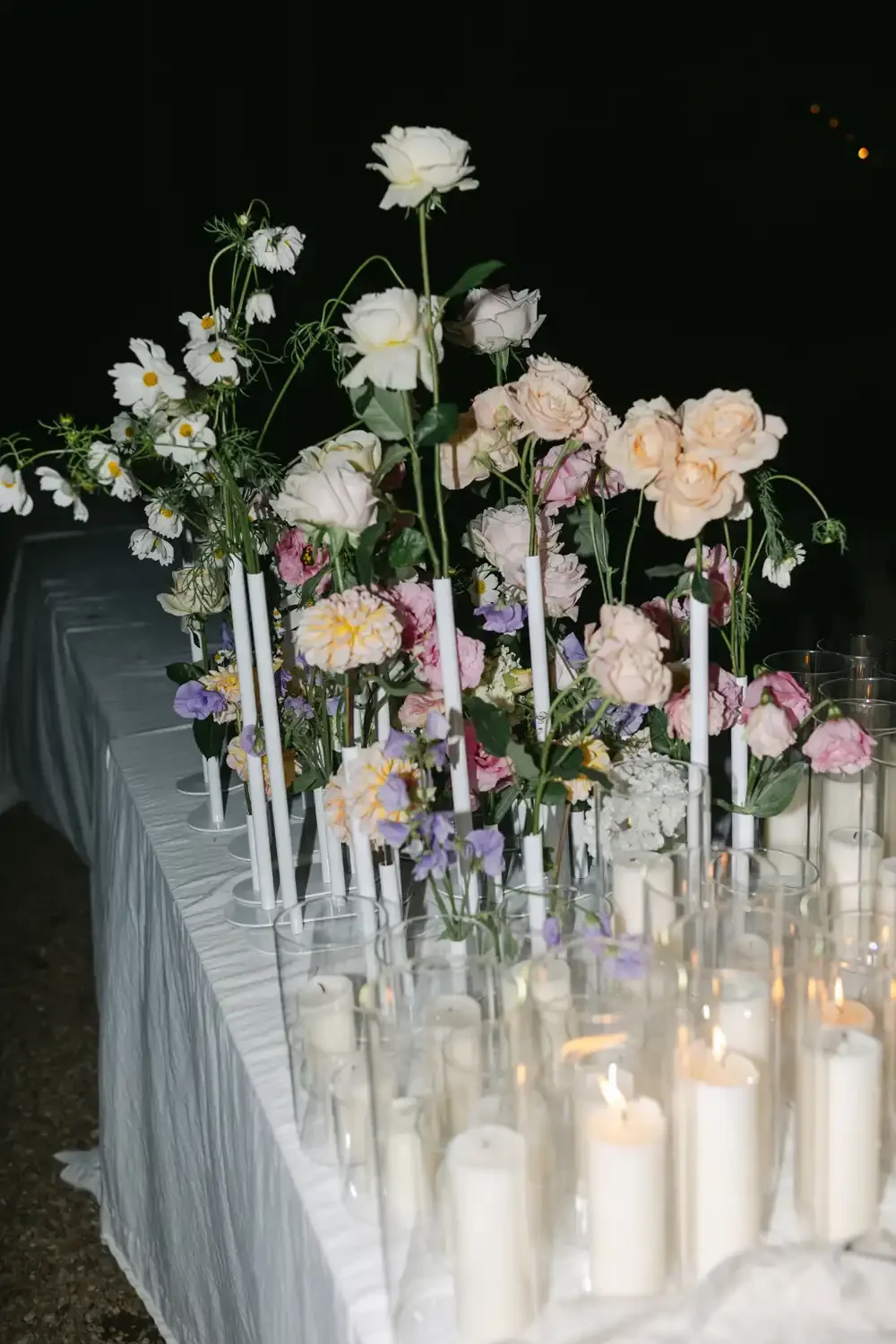 A floral centerpiece with white, pink, and purple flowers on a table decorated with candles in glass holders, set against a dark background.