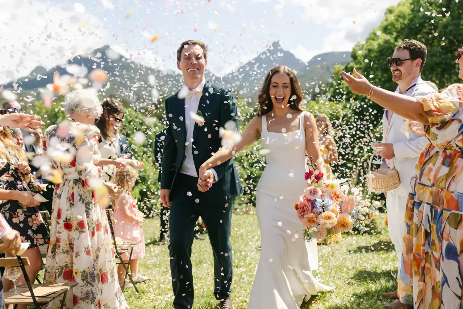 A newly married couple holding hands and smiling as they walk through a confetti shower outdoors at a wedding reception. The bride is wearing a white dress and holding a floral bouquet; the groom is in a dark suit with a white bow tie. Guests around 