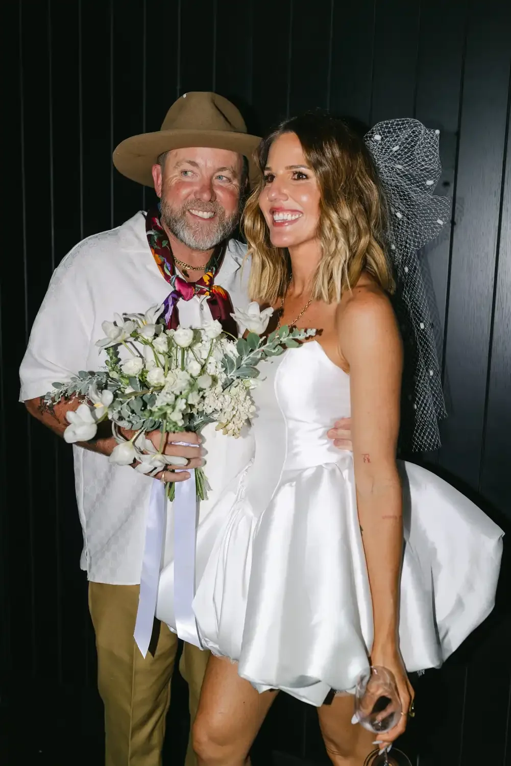 A man and woman dressed in wedding attire, smiling and holding a bouquet of white flowers, standing against a black wood-paneled wall.