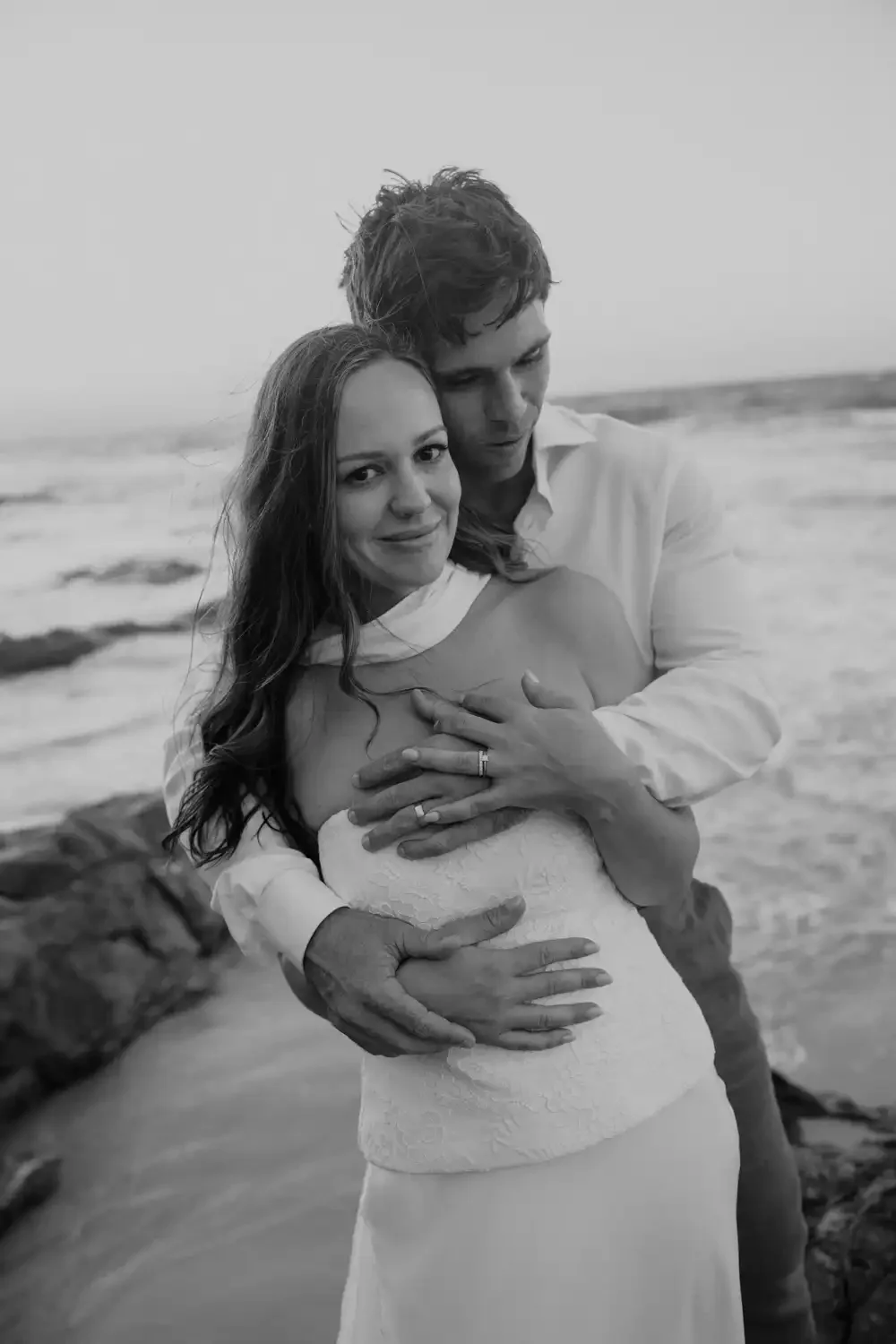 Black and white photo of a couple embracing on the beach. The woman is facing the camera with a slight smile, while the man is looking down at her. Their hands, with wedding rings, are intertwined on her chest. The ocean and rocks are in the backgrou