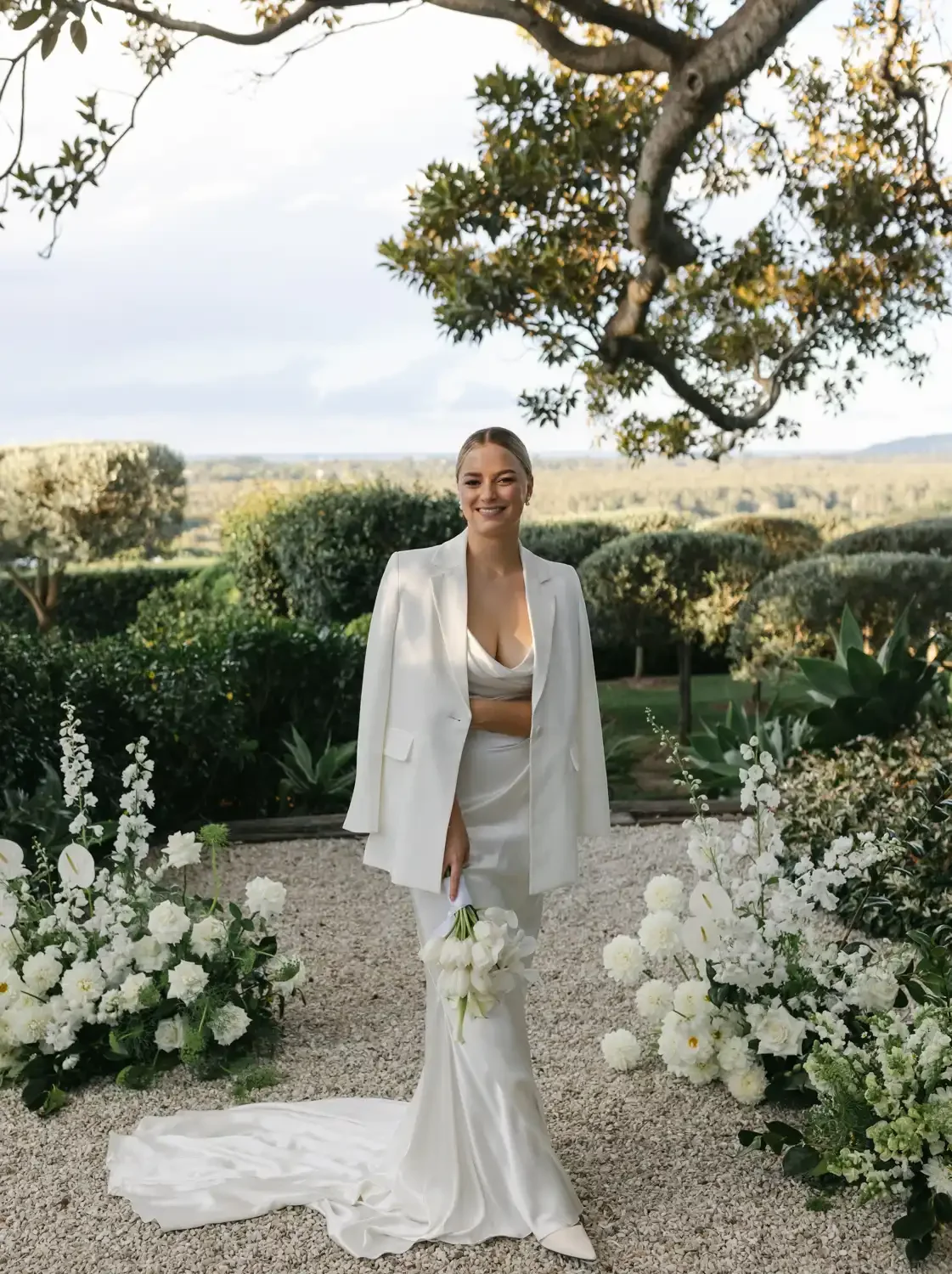 A woman in a white dress and blazer holding a bouquet of white flowers, standing outdoors in a garden with white flowers and green foliage, and a tree overhead, during daytime.