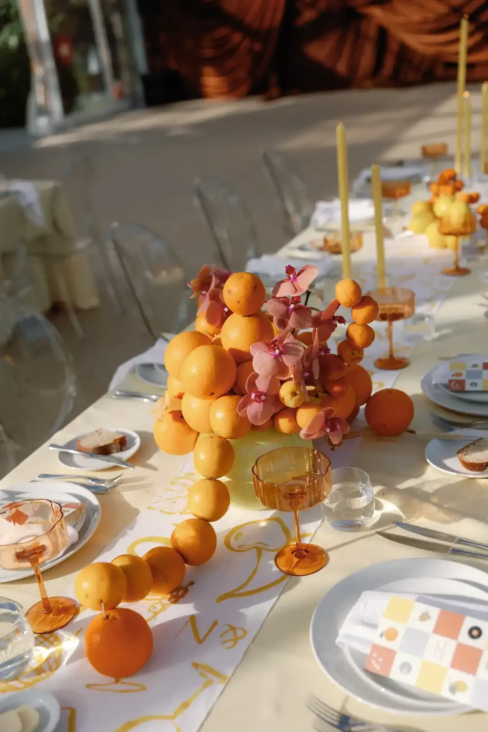 A decorated outdoor dining table with a floral centerpiece made of oranges and pink flowers, set with plates, glasses, and utensils for a celebration.