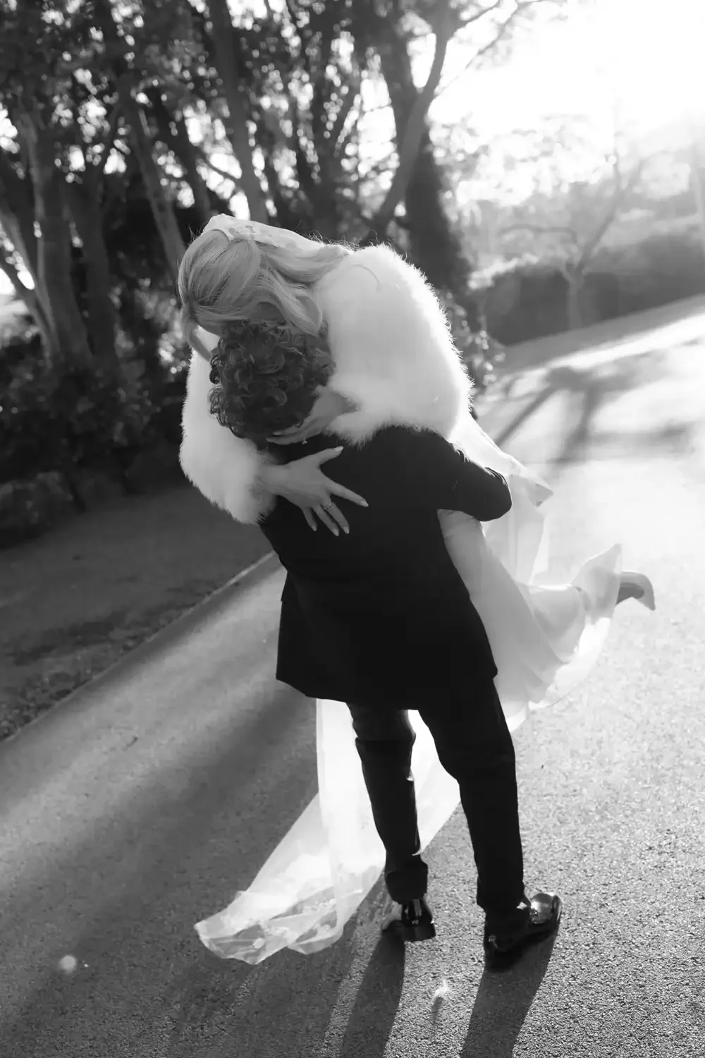 A person in a tuxedo lifting and holding a woman in a wedding dress in an outdoor setting, with trees and sunlight in the background, in black and white.