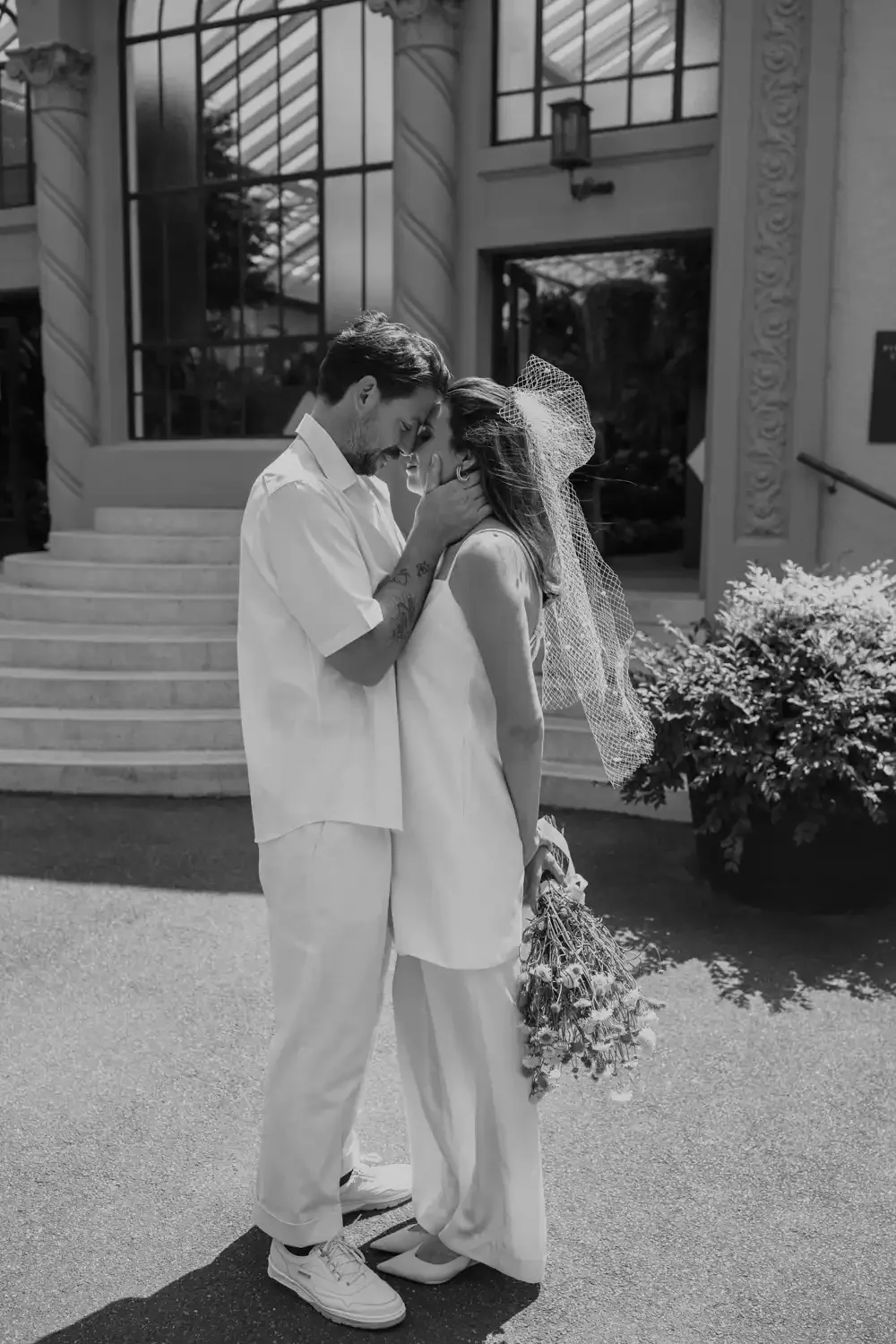 A black and white photo of a couple embracing outside a building. The man has his forehead against the woman's, holding her face with both hands. The woman is holding a bouquet of flowers, and she is wearing a veil. Both are dressed in light-colored 