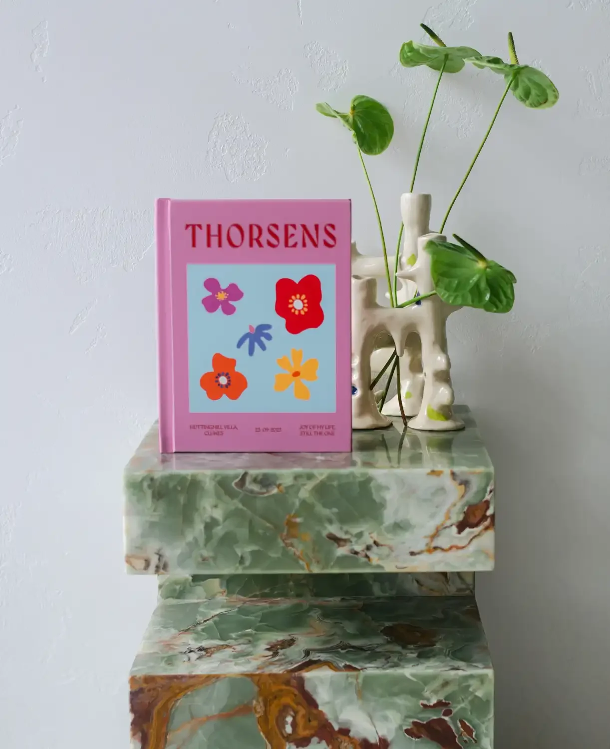 Pink book titled 'Thorsens' with colorful flower illustrations, placed on a marble table with a ceramic vase holding green leafy plants against a white textured wall.
