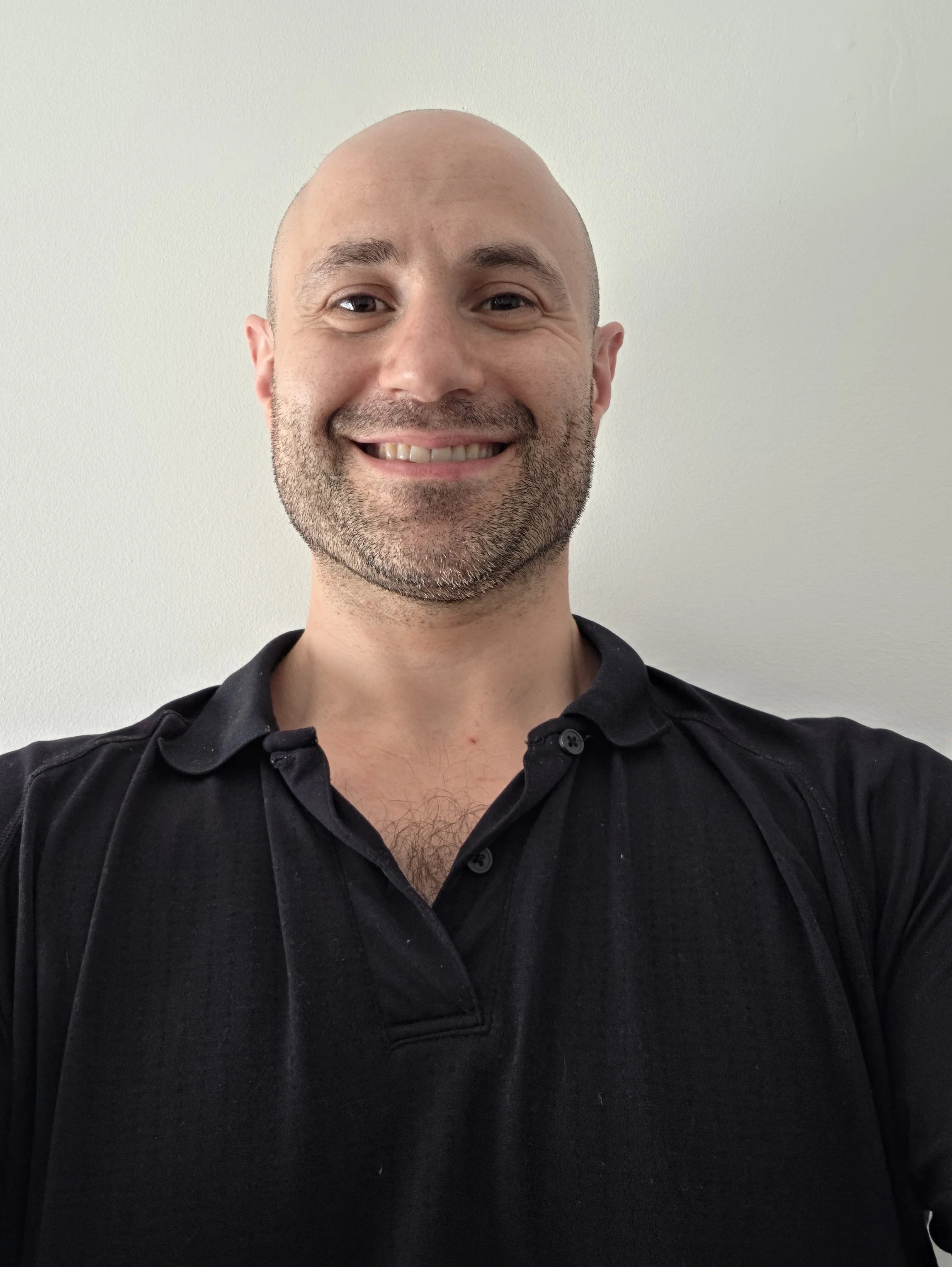 Smiling bald man with a short beard wearing a black collared shirt in a close-up portrait.
