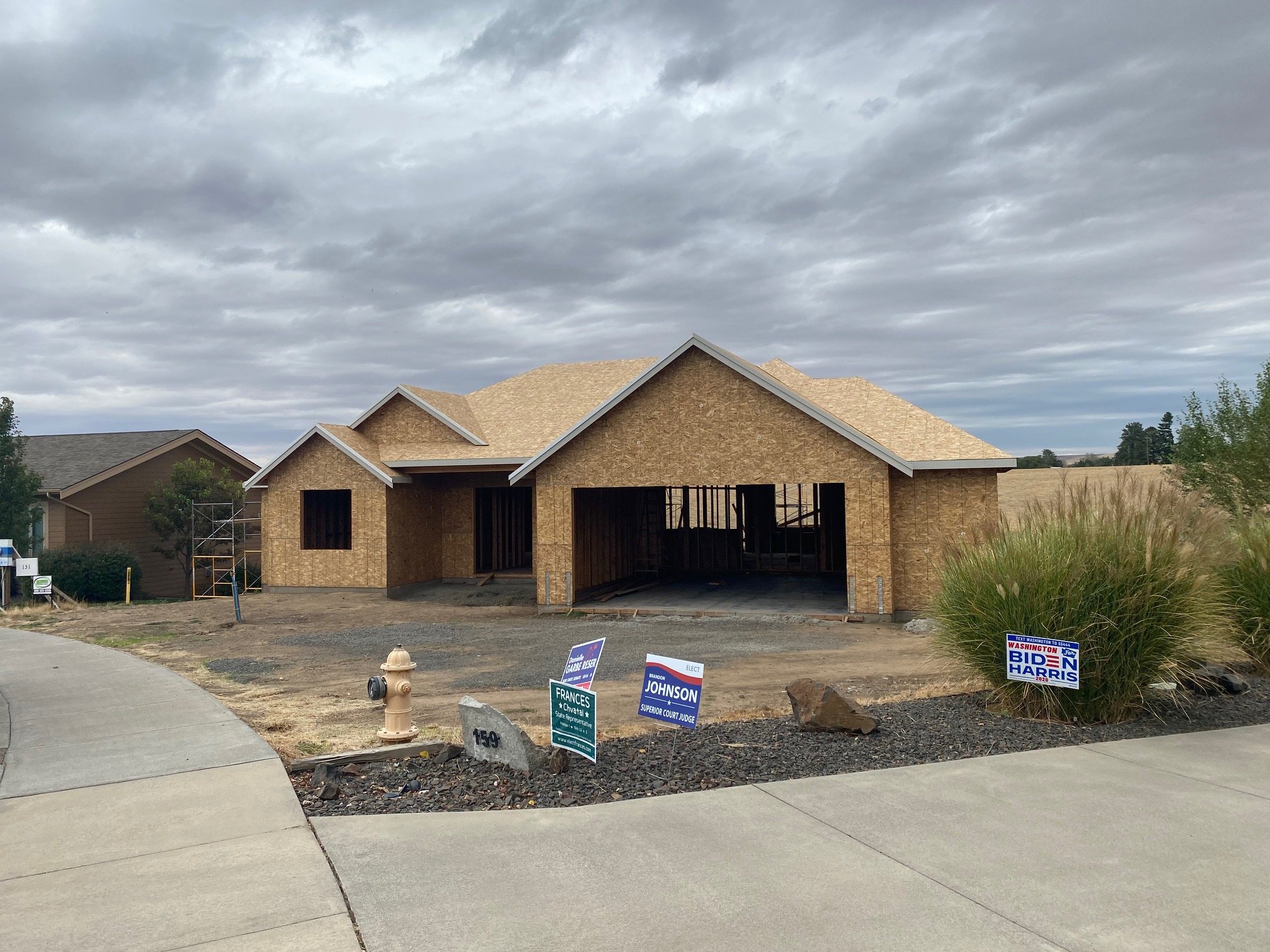 A house under construction with wooden framing and no exterior walls, located in a suburban area. There are campaign signs for political candidates in the yard, including Biden-Harris, Johnson, and Frances.