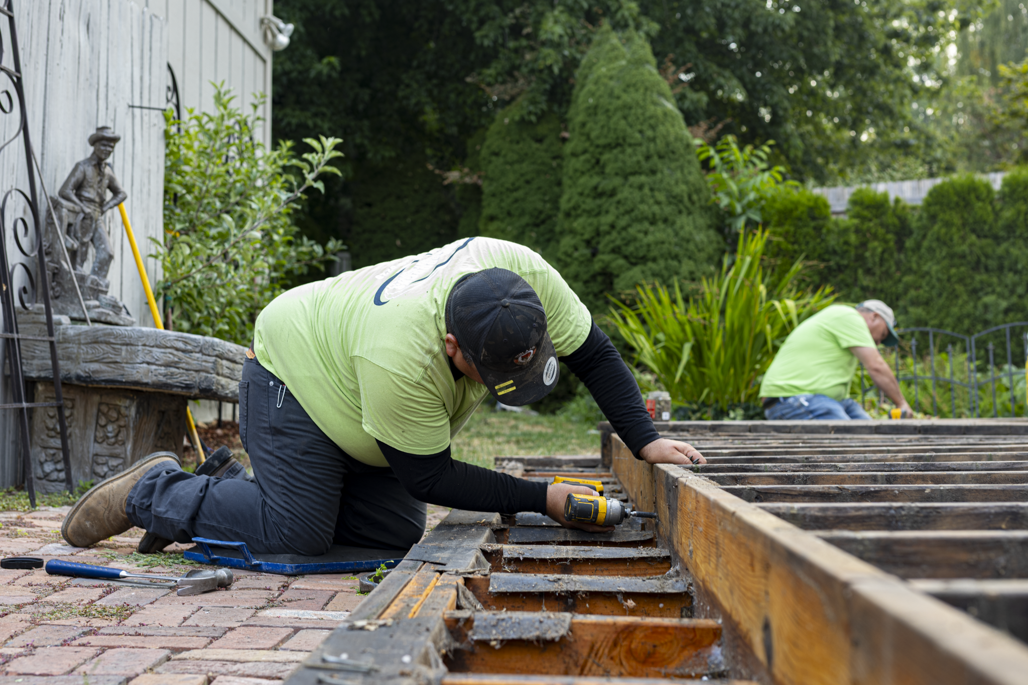 Two workers crouched down, working on a wooden garden bed outdoors. One worker is using a drill, and there are tools around them. The background features lush green trees and shrubs.