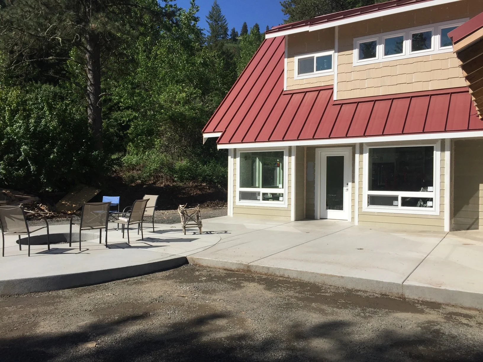 Newly constructed house with beige siding, red metal roof, and large windows. Outdoor patio area with chairs, a small decorative wooden horse, surrounded by trees and greenery.