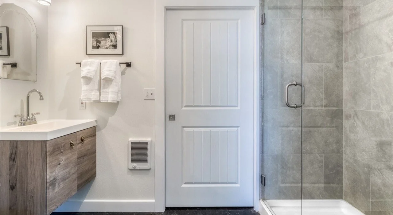 Bathroom with a wooden vanity, white sink, mirror, towel rack with white towels, framed black and white photograph, closed white door, and a walk-in shower with a glass door and gray tiled walls.