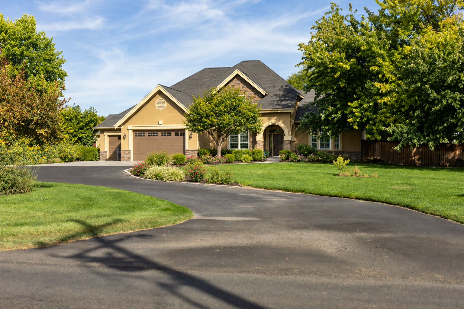 A large, well-maintained house with a curved driveway, surrounded by lush green lawn and trees, under a blue sky with some clouds.